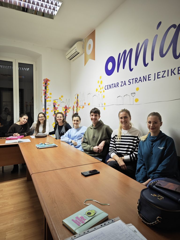 Eight students sit along a long wooden table in a bright classroom, smiling at the camera with colorful wall art behind them.