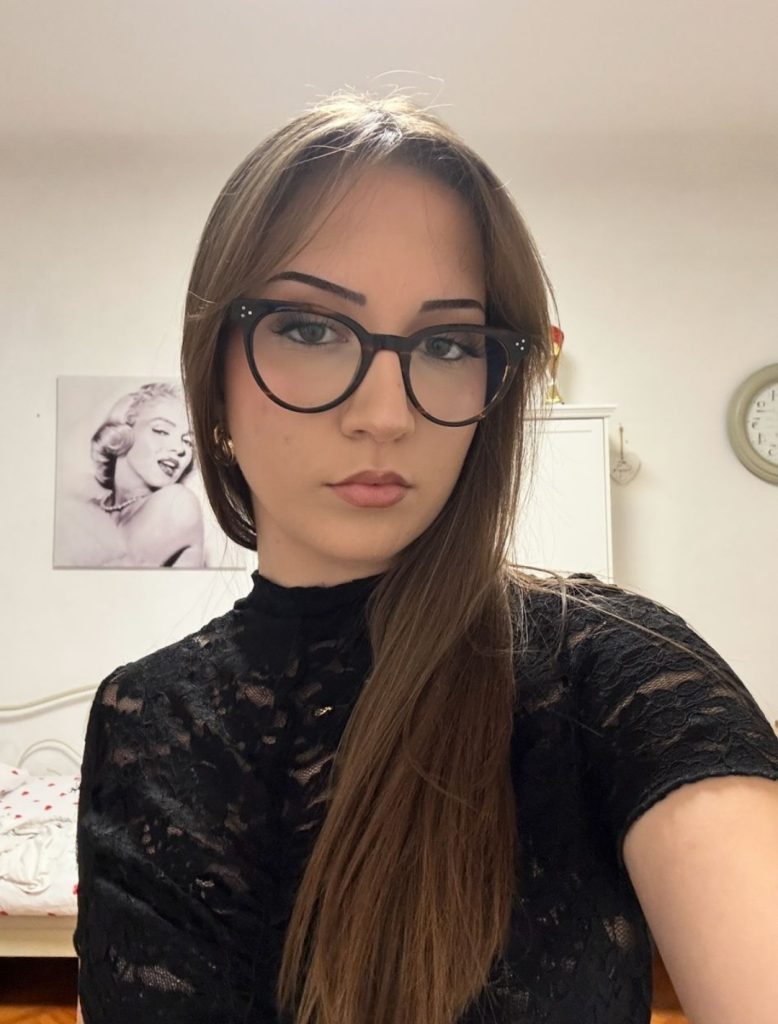 Young woman with long brown hair and black-rimmed glasses, wearing a black lace top, in a bedroom with a Marilyn Monroe poster on the wall.
